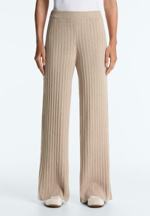 Beige bordázott kötött széles szárú nadrág fehér tornacipővel viselve, álló emberen, deréktól lábig látható.