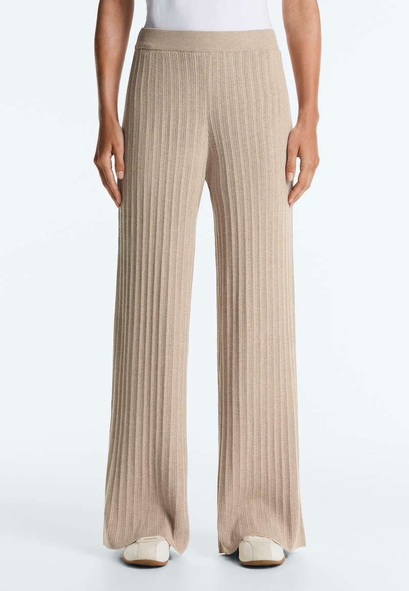 Beżowe szerokie spodnie z prążkowanego dzianiny, noszone z białymi trampkami na stojącym osobie, widoczne od talii do stóp.