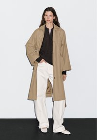 Beige lange jas met oversized pasvorm, gedragen over een donkere gebreide trui, gecombineerd met brede witte broeken en lichtgekleurde instappers.