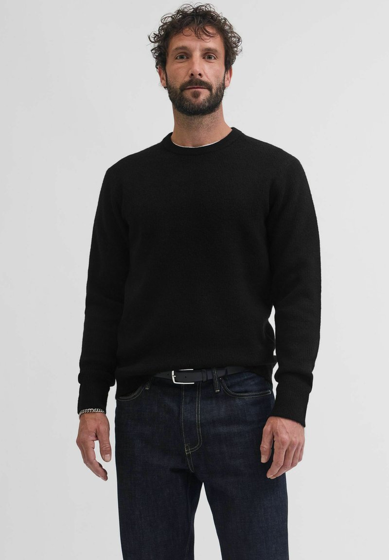 Maglione nero in maglia con scollatura tonda e polsini a costine, indossato sopra jeans blu scuro. Tessuto liscio, vestibilità comoda, design minimale.