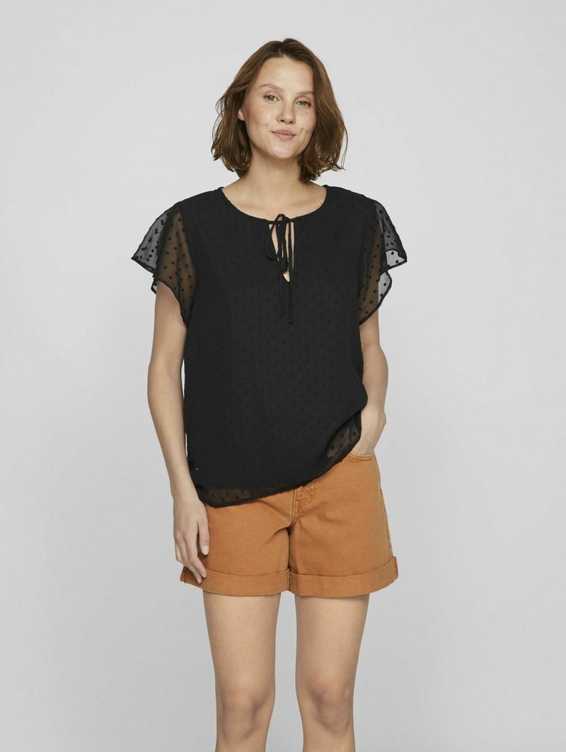 Svart topp med korte, gjennomsiktige ermer med prikker og knyting i halsen, sammen med oransje shorts med rullet kant. Tekstilet er lettvektig og har et uformelt design.