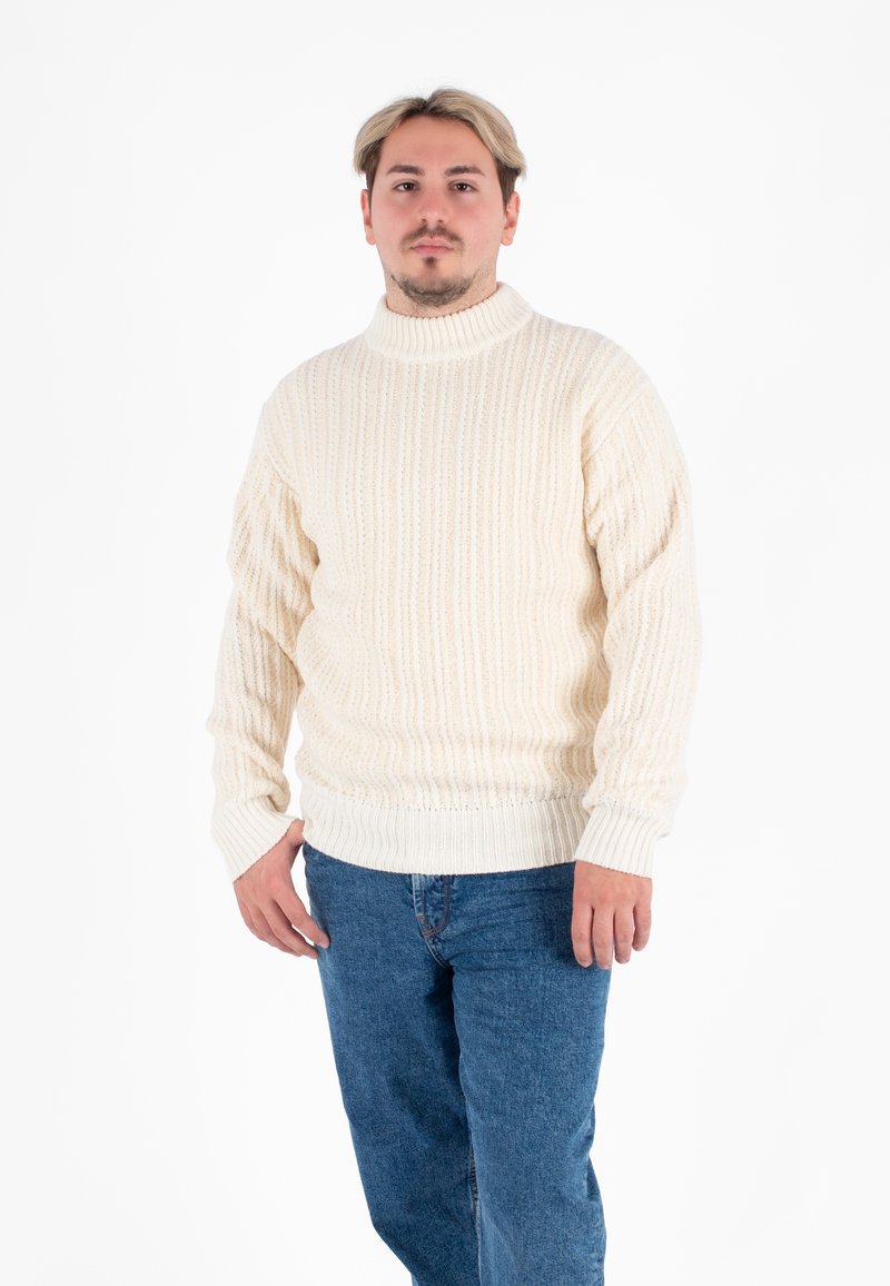Cremfarbener Strickpullover mit gerippter Textur und hohem Kragen, kombiniert mit blauen Jeans. Das Modell steht vor einem weißen Hintergrund.