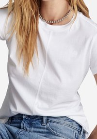 T-shirt en coton blanc à manches courtes, avec un logo subtil et des coutures contrastantes. Associé à un jean en denim bleu. Collier en perles argentées.