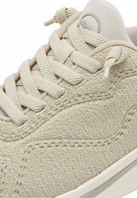 Gros plan sur une basket en tissu beige avec des lacets épais et texturés et une languette rembourrée portant un petit logo circulaire.