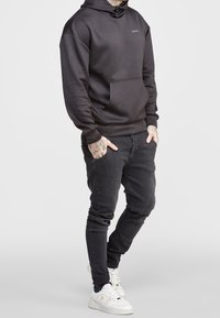 Sudadera negra con un bolsillo frontal, hecha de una tela suave, con un logo minimalista. Combinada con jeans oscuros y zapatillas blancas.