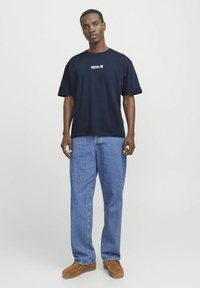 Mørkeblå bomulds T-shirt med "SEOUL" grafik, parret med lyseblå denimjeans og brune ruskindsko. Modellen står op af en hvid baggrund.