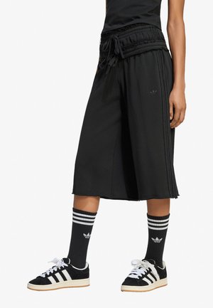 Persona che indossa pantaloni neri a gamba larga e corta, calzini alti neri Adidas con strisce bianche e sneakers nere Adidas con strisce bianche.