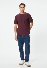 Maroonowa, krótko-sleeve koszulka z bawełny, z okrągłym dekoltem, w połączeniu z niebieskimi dżinsami i białymi trampkami z brązowymi akcentami. Postawa stojąca.