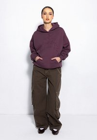 Fioletowa bluza z kapturem z wytłoczonym napisem "LOCAL HEROES", w połączeniu z oversize'owymi brązowymi spodniami cargo z bocznymi kieszeniami i luźnym krojem.