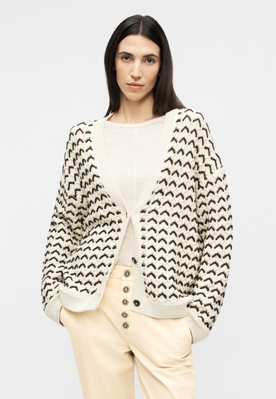 Γυναίκα με ίσια σκούρα μαλλιά, φοράει ζακέτα με κρεμ και μαύρο μοτίβο πάνω από λευκό μπλουζάκι και μπεζ παντελόνι ψηλόμεσο με διακοσμητικά κουμπιά.