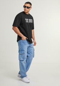 Fekete, oversized póló "TRUST ISSUES" felirattal, világoskék cargo farmernadrág oldalzsebekkel és fehér sneaker cipővel. Textúrált anyag, laza szabás.