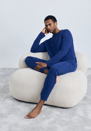 Ensemble de haut à manches longues et de pantalon bleu marine en tissu doux, porté par une personne assise sur une chaise beige ronde et texturée, sur un tapis gris.