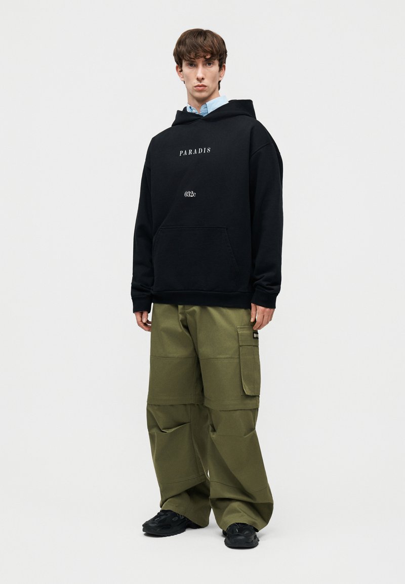Joven con sudadera con capucha negra con el texto "PARADIS 032e", pantalones cargo verde oliva y zapatillas negras, de pie contra un fondo liso.