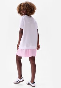 Witte oversized t-shirt met korte mouwen, gelaagd over een roze gestreepte plissérok, gecombineerd met witte sneakers met zwarte accenten.