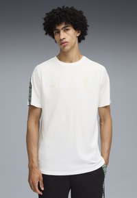 Weißes T-Shirt mit kurzen Ärmeln, Rundhalsausschnitt, grünen Musterakzenten an den Ärmeln und goldenen Logos von Puma und "KING".