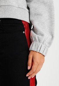 Mano appoggiata su pantaloni neri con striscia di paillettes rosse, indossa una felpa grigia chiara con polsini elastici.