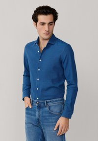 Muž v priliehavých modrých košeli na gombíky zastrčenej do svetlomodrých džínsov, stojaci s jednou rukou v vrecku pred jednoduchým pozadím.