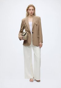 Kvinde iført en tanfarvet blazer, hvid top, brede hvide bukser og sandaler, der holder en tekstureret beige clutch mod en ensfarvet baggrund.