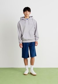 Sweatshirt à capuche gris avec poche frontale, associé à un short en denim bleu, des chaussettes blanches et des baskets blanches sur un sol vert.