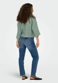 Top verde a maniche 3/4, con texture, abbinato a jeans slim-fit blu scuro e scarpe nere flat, vista del modello da dietro.
