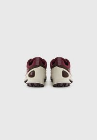 Scarpe sportive con tomaia in tessuto color marrone scuro, dettagli sintetici beige e suola esterna marrone resistente con trazione. Presentano un tallone rinforzato e un collarino imbottito.