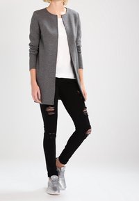 Cappotto lungo grigio texturizzato con scollo rotondo, indossato su una maglietta bianca, abbinato a jeans neri strappati e sneaker argento.