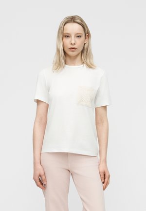 Donna bionda che indossa un top bianco a maniche corte con una tasca color crema testurizzata e pantaloni rosa chiaro, in piedi davanti a uno sfondo chiaro e uniforme.