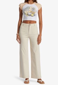 Frau trägt ein weißes Crop-Top mit tropischem Fruchtmuster, beigefarbene hochgeschnittene, weite Hosen, ein Muschelarmband und weiße geflochtene Sandalen.