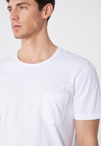 Camiseta corta de manga blanca en tejido ligero con escote redondeado y un bolsillo parche en el lado izquierdo del pecho. Textura suave.