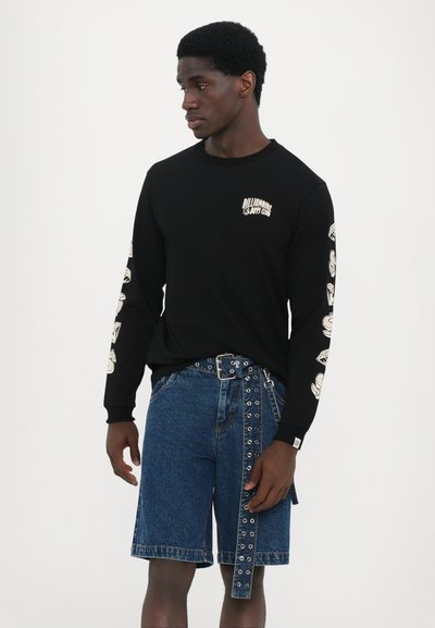 Sort langærmet skjorte med logo på brystet og mønstrede designs på ærmerne, kombineret med blå denimshorts med unikke bælte detaljer.
