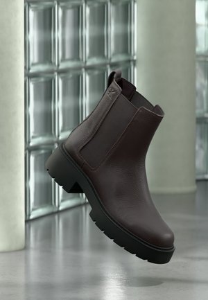 Dark brown leather Chelsea boot with elastic side panels and chunky black sole, shown floating inside a modern room with glass block wall.