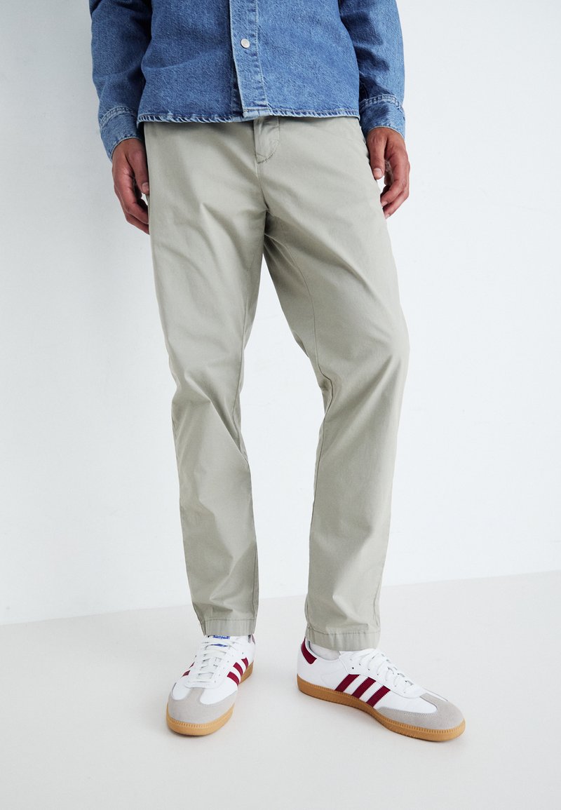 Pantaloni di cotone beige chiaro abbinati a una camicia di jeans blu e sneaker bianche con strisce rosse e suole di gomma, esposti in posizione verticale.