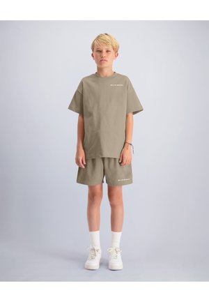 Jongen die een taupekleurig oversized t-shirt en bijpassende korte broek draagt, witte sokken en witte sneakers, staand tegen een effen lichtgrijze achtergrond.