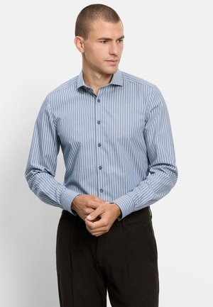 Mann trägt ein blau-weiß gestreiftes langärmeliges Hemd mit Knöpfen und schwarze Hose, schaut nach rechts mit verschränkten Händen.