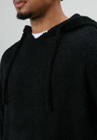 Schwarzer Hoodie aus Plüschstoff mit strukturierter Oberfläche, Kordelzugkapuze und lockerem Schnitt. Minimalistisches Design ohne Muster.