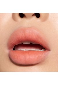 Gros plan sur des lèvres d'un ton pêche-rose mat. Texture lisse, forme légèrement pulpeuse, légèrement entrouvertes, laissant entrevoir des dents blanches.