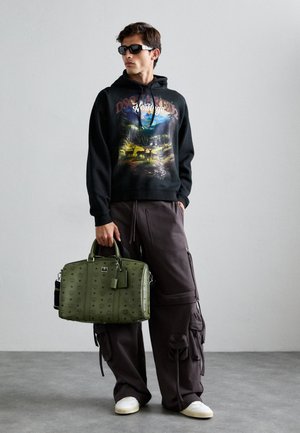 Black hoodie with graphic print, dark cargo pants, and green textured bag. Features dual handles, metal accents, and logo detail. White sneakers.