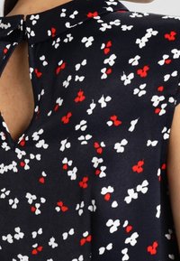 Blusa blu navy con colletto, decorata con un motivo floreale di fiori rossi e bianchi. Tessuto morbido, maniche corte a cuffia e dettaglio con apertura a goccia sul collo.