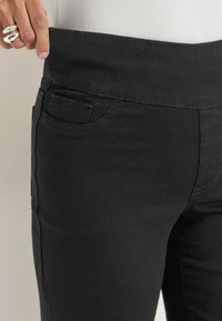 Käsi, joka vetää mustia venyviä korkeavyötäröisiä housuja, mikä korostaa istuvuutta ja mukavuutta, sormissa näkyviä renkaita.