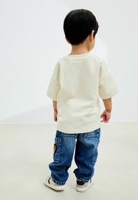 T-shirt oversized color crema con maniche corte, abbinato a jeans in denim blu a vestibilità ampia con tasche laterali e dettaglio a patch circolare.