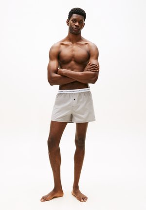 Uomo senza maglietta in piedi a piedi nudi con le braccia incrociate, indossa boxer grigio chiaro con una cintura brandizzata.