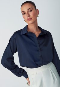 Blusa in raso blu navy con colletto classico, maniche lunghe e chiusura con bottoni; presenta una texture liscia e un taglio morbido.