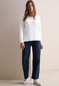 Witte longsleeve top met een V-halsontwerp, gecombineerd met donkerblauwe recht gesneden jeans en witte sneakers op een neutrale achtergrond.