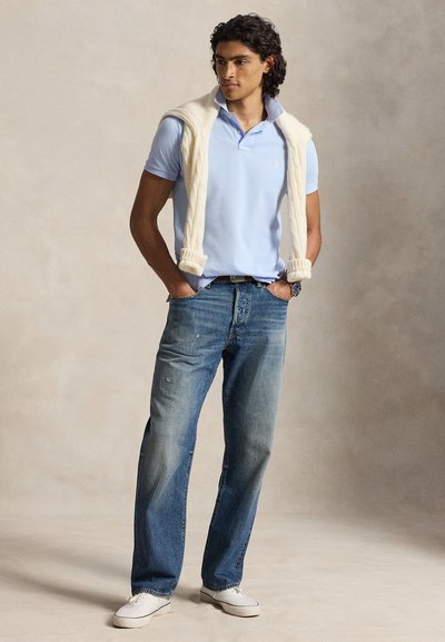 Jeune homme portant un polo bleu clair avec un pull crème drapé sur les épaules, jean bleu, baskets blanches, debout les mains dans les poches.