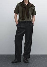 Polo à manches courtes vert foncé, pull marron porté sur les épaules, pantalon large noir et chaussures noires avec lacets marron.