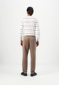 Uomo di spalle che indossa una camicia bianca a maniche lunghe con strisce nere e rosse, pantaloni in velluto a coste marroni e scarpe nere su sfondo bianco.