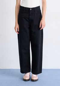 Black wide-leg jeans with a high waist, featuring front pockets and a button closure, paired with light pink shoes on a blue floor.