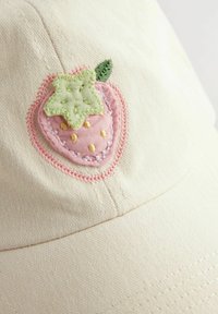 Gorra de béisbol color crema con un diseño de fresa bordado, con acentos en rosa y verde y detalles de semillas amarillas. Fabricada con tejido texturizado.