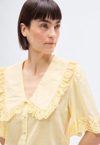 Femme aux cheveux courts et foncés portant un chemisier jaune pâle avec un large col à volants et des détails en dentelle ajourée sur les manches et le col.