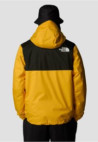 Gelbe und schwarze, wasserdichte Kapuzenjacke mit elastischen Bündchen und einem Logo auf dem Rücken. Begleitet von einem schwarzen Fischerhut.